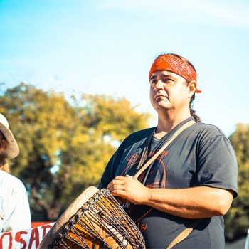 Andrew Ecker, with a red bandana around his head, looks sternly in the distance while holding a drum strapped over his shoulder.